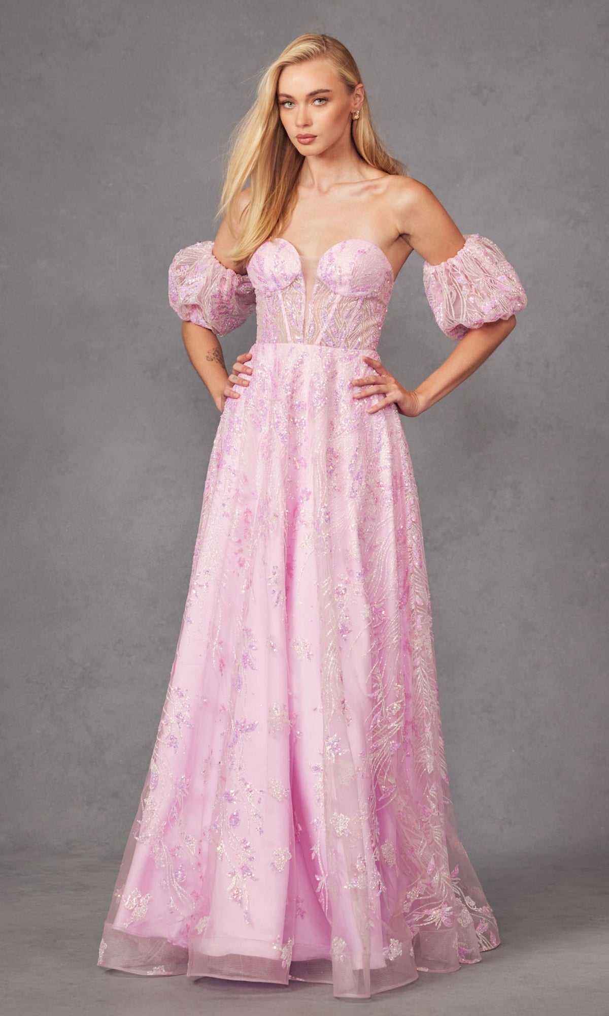 A woman in the Puff-Sleeve Strapless Long Glitter Prom Dress 2404, featuring light pink fabric, sheer puff sleeves, and floral embellishments, stands against a plain gray background.